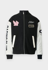 Αμάνικο μπουφάν με φερμουάρ σε μαύρο και άσπρο χρώμα με λογότυπα "Hilfiger" και "Championship Team 1985", σήμα με φτερά και τα αρχικά "TH" καθώς και το κείμενο "Racing Club" στα μανίκια.