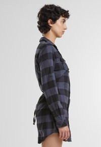 Abito camicia in flanella a quadri blu e neri con maniche lunghe, chiusura a bottoni sul davanti, fiocchi laterali e due taschini sul petto, mostrato in vista laterale.