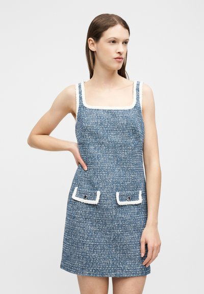 Jeune femme debout, main sur la hanche, portant une robe sans manches en tweed bleu avec des bordures blanches et deux poches avant, sur fond uni.