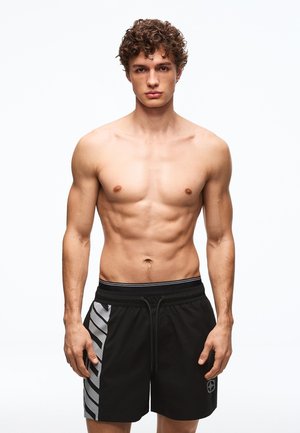 Jeune homme aux cheveux bouclés, debout face à l'avant, torse nu, portant un short de sport noir avec des rayures diagonales grises sur le côté gauche.