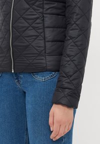 Svart quiltet jakke med glidelås foran, glatt tekstur og en tettsittende design. Kombinert med blå denimbukser.