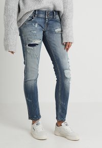 Jean skinny bleu déchiré avec des trous et des patchs délavés, associé à un pull en tricot gris clair et des baskets blanches.