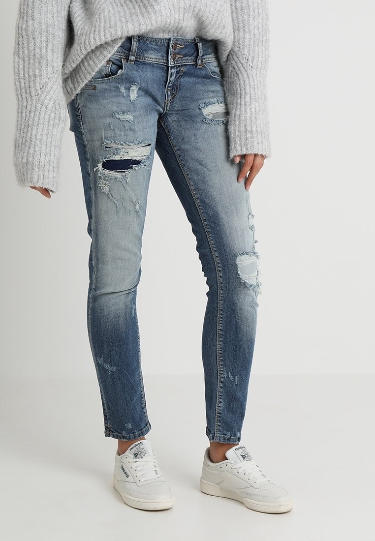 Jean skinny bleu déchiré avec des trous et des patchs délavés, associé à un pull en tricot gris clair et des baskets blanches.