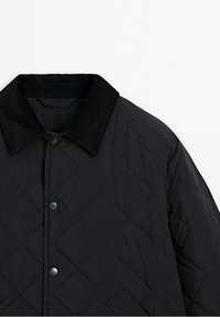 Musta tikattu takki, jossa on pikeerinen kaulus. Sisältää pikalukitusnapit ja teksturoidun kangaspinnan. Yläosan vinosta näkökulmasta.