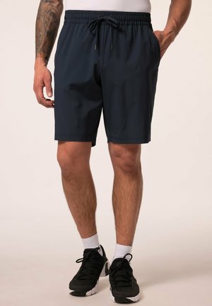 Mann, der schwarze Shorts mit Kordelzug, weiße Socken und schwarze Sportschuhe trägt, steht mit einer Hand in der Tasche und sichtbar tätowiertem Arm.
