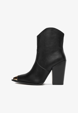 Czarny skórzany botek za kostkę z wysokim słupkowym obcasem i spiczastym noskiem, z złotym metalowym akcentem na czubku. Gładka tekstura.