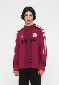 Jauns vīrietis ar melniem cirtainiem matiem, ģērbies bordo Adidas džemperī ar baltām svītrām uz piedurknēm un melnās biksēs, fona ir balts.