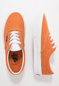 Baskets en toile orange avec semelles en caoutchouc blanc et lacets, dotées d'un col rembourré et d'une étiquette de marque noire. La texture est lisse et durable.