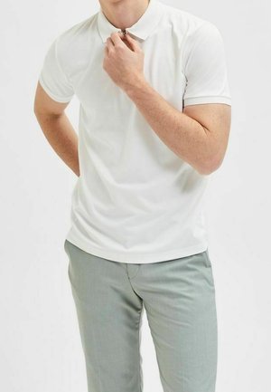 Mann trägt ein weißes Poloshirt mit kurzen Ärmeln und hellgraue Hose, hält mit einer Hand den Reißverschluss des Shirts in der Nähe des Kragens.