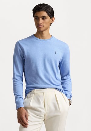 Lichtblauwe crewneck trui gemaakt van een gladde stof, met lange mouwen en een klein donker logo aan de linkerkant. Draag het met crème kleurige broek.