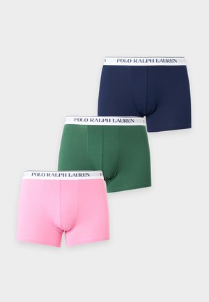 Mladý muž s tvarovanou postavou v zelených boxer nohavičkách s bielym pásikom s označením "Polo Ralph Lauren", stojaci na jednoduchom pozadí.