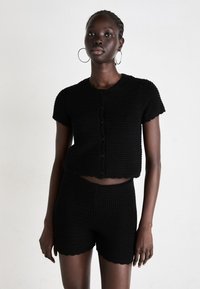 Crni pleteni crop top s kratkim rukavima i dugmadi, u kombinaciji s odgovarajućim visokim hlačama, koje imaju valovite rubove.