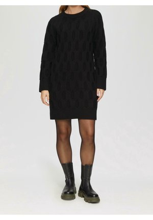 Vrouw draagt een zwarte kabelgebreide sweaterjurk, doorschijnende zwarte panty's en stevige zwarte enkellaarzen, staand tegen een effen achtergrond.