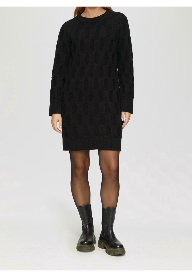 Femme portant une robe-pull en maille torsadée noire, collants noirs transparents et bottines noires épaisses, debout sur un fond uni.