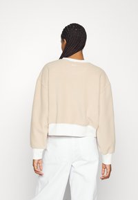 Beige kort sweatshirt med en mjuk textur, vita ribbade ärmslut och en subtil logotyp på baksidan. Bärs över vita byxor.