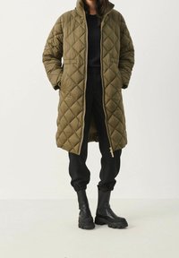 Oliven grøn quiltet puffjakke med høj krave og frontlynlås, parret med sorte joggers og chunky sorte støvler, vist mod en lys baggrund.