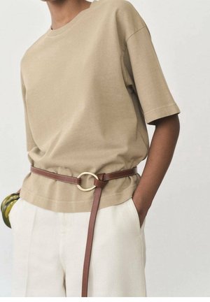 Personne portant un haut beige à manches courtes rentré dans un pantalon blanc avec une ceinture en cuir marron ornée d'une grande boucle circulaire en or.