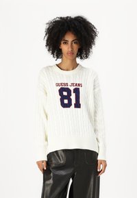 Maglione bianco a maglia cable con scollo rotondo, caratterizzato da una grafica rossa e blu navy "GUESS JEANS 81" sulla parte anteriore, abbinato a pantaloni neri.