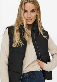 Gilet rembourré noir avec col haut et fermeture éclair, superposé à un pull clair. Porté avec un jean foncé, présentant un look décontracté.