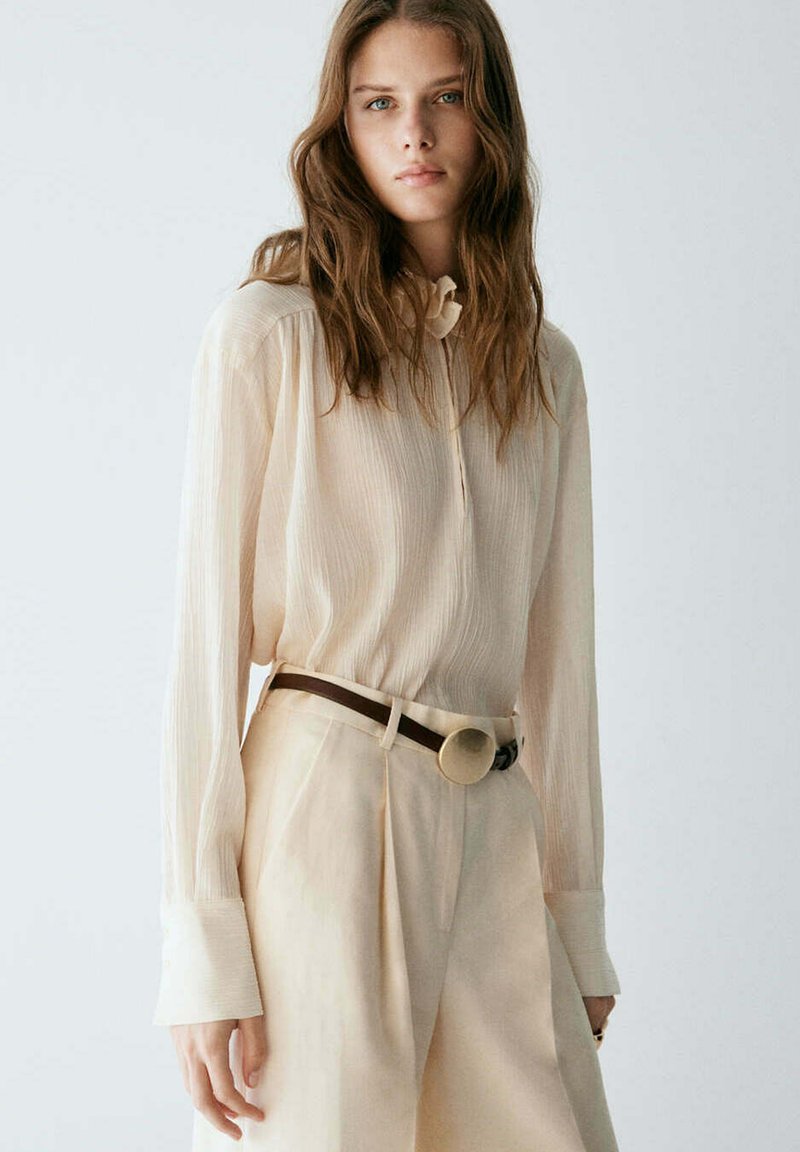 Femme aux longs cheveux bruns portant un chemisier léger beige transparent rentré dans un pantalon beige taille haute avec une ceinture foncée et une boucle ronde.