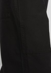 Fekete szövetnadrág, egyenes szabással, varrás részletekkel és oldalsó zsebbel. Az anyag sima és enyhén texturált felületű.