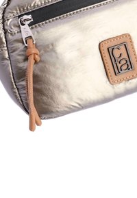 Neceser cosmético plateado metálico con acabado texturizado, que presenta una cremallera negra y acentos de cuero marrón, incluyendo un parche con el logo y un tirador de cremallera.