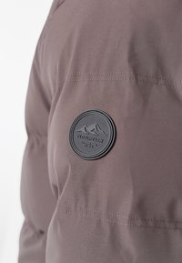 Brązowa kurtka na zewnątrz o gładkiej fakturze, z okrągłą czarną gumową naszywką z logo i widocznymi detalami szwów.