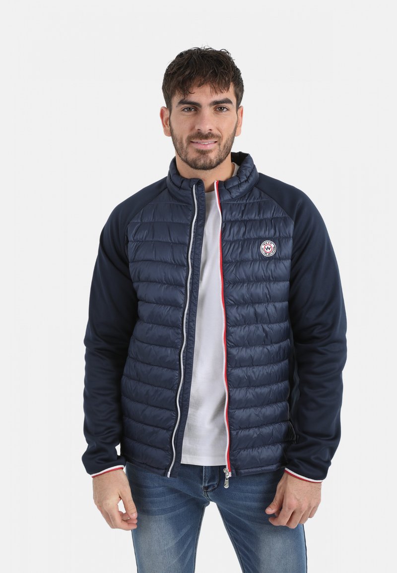 Veste bleu marine avec un devant matelassé, fermeture éclair blanche et accents rouges. Présente des manches longues et un col montant. Coupe décontractée, textures contrastantes.
