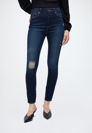Femme portant un jean skinny bleu foncé avec un détail usé au genou et des mules noires à talons, debout sur un fond blanc.