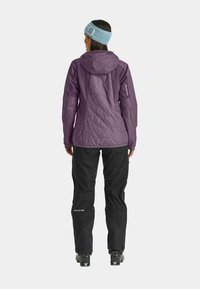 Purple insulated jacket with quilted texture, hood, and logo detail on sleeve, paired with black waterproof pants and ski boots.