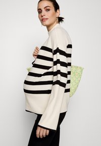 Pullover crema con righe nere, vestibilità ampia, maniche lunghe e scollo rotondo. Accostato a una borsa a tracolla verde e gialla con texture.