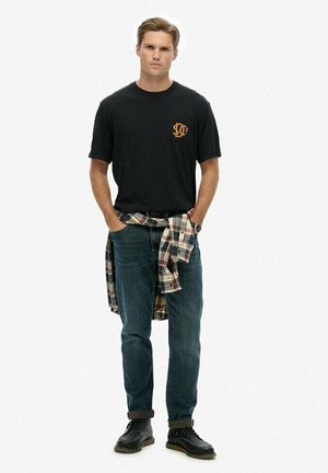 Zwart T-shirt met korte mouwen en oranje grafiek, blauwe jeans en tan laarzen. Een flanellen overhemd is om de taille geknoopt. Neutrale achtergrond.