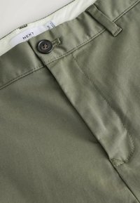 Pantalon slim vert olive avec fermeture à bouton, passants pour ceinture et étiquette intérieure visible indiquant "NEXT."