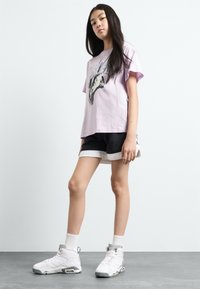 Lys lilla bomull t-skjorte med grafisk trykk, kombinert med svart og hvit treningsshorts. Modellen har på seg hvite høye sneakers.