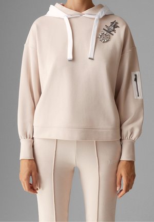 Kobieta w beżowej bluzie z kapturem ozdobionej broszkami z kryształków i dopasowanych spodniach, stojąca na szarym tle.