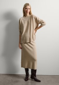 Suéter de punto beige con corte holgado y dobladillo acanalado, combinado con una falda midi a juego. La modelo lleva botas de charol burdeos hasta la rodilla.