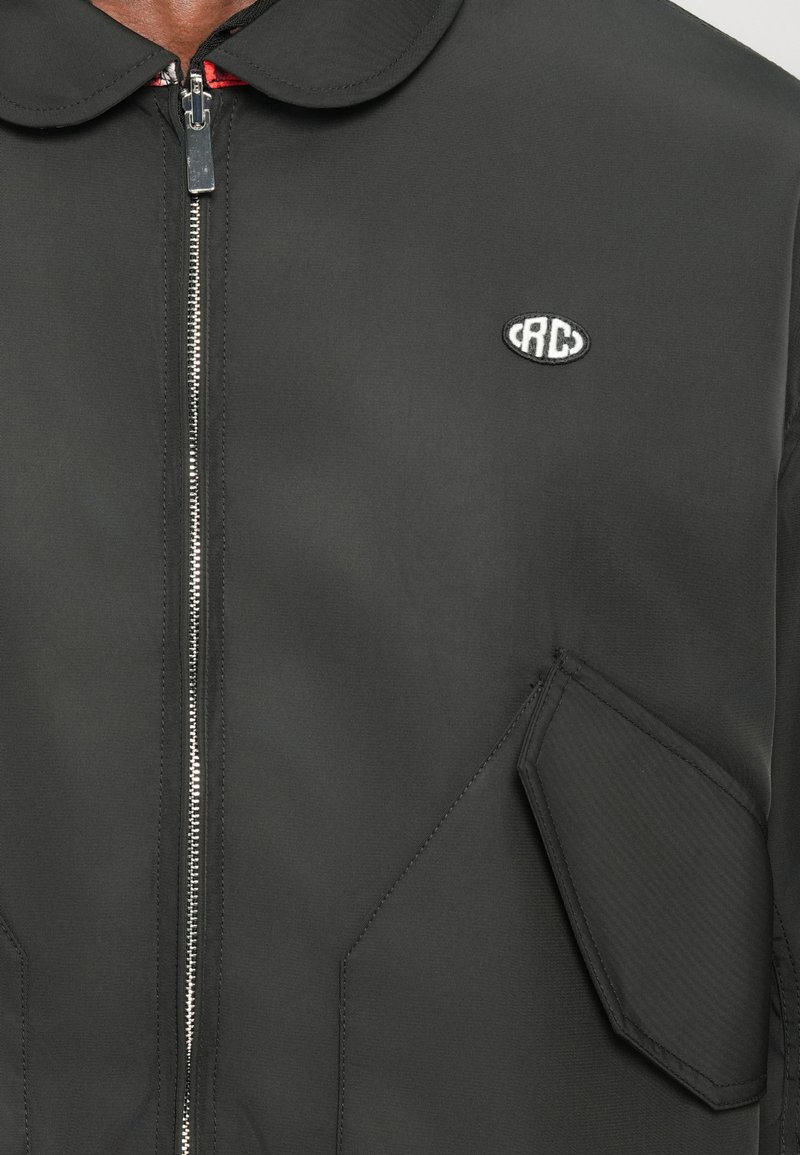 Veste noire avec fermeture éclair sur le devant, col arrondi et patch logo. Le tissu a une texture lisse, une coupe ajustée et des poches inclinées.