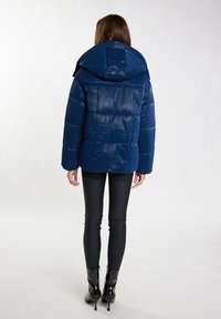 Marineblå pufferjakke med quiltet design, overdimensioneret hætte og en glansfuld tekstur. Kombineret med sorte tætsiddende bukser og glansfulde ankelstøvler.