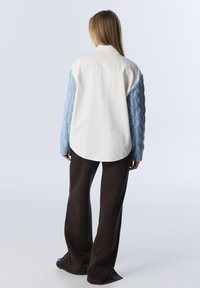 Chemise blanche avec manches en maille torsadée bleu clair, ourlet arrondi, portée avec un pantalon large marron. Design simple, manches texturées, sans motifs visibles.