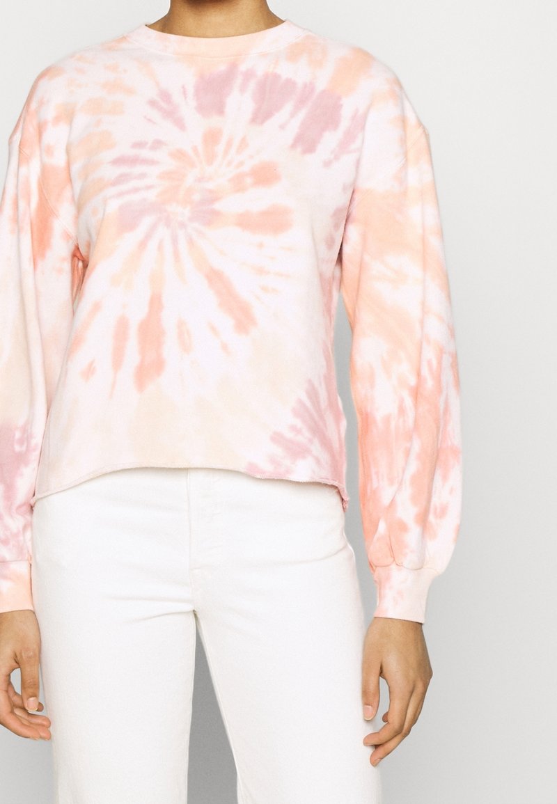 Felpa tie-dye nei toni pesca e rosa, collo a giro, maniche lunghe, tessuto morbido, orlo corto con una vestibilità rilassata. Abbinata a pantaloni bianchi.