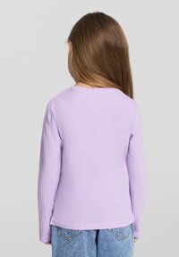 Fioletowa koszula z długim rękawem i okrągłym dekoltem, wykonana z miękkiego materiału. Ma gładką fakturę i luźny krój, zestawiona z dżinsowymi szortami.