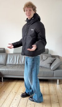 Čierna nadýchaná bunda s kapucňou, vybavená predným zipsom a logom, v kombinácii s voľnými modrými džínsy a čiernymi topánkami na drevenej podlahe.