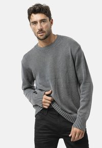 Pull en tricot gris avec un col et des poignets côtelés, présentant une coupe décontractée et une texture subtile. Porté par-dessus un pantalon noir.