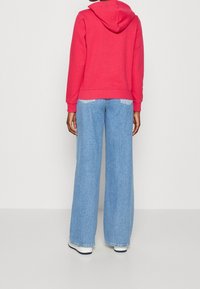 Persona che indossa un felpa rossa, jeans a gamba larga di colore blu chiaro e sneakers bianche, in piedi di spalle contro uno sfondo bianco uniforme.