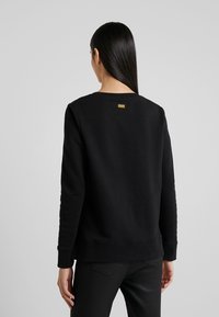 Černá mikina s kulatým výstřihem, dlouhými rukávy a strukturovanými pletenými manžetami. Na horní části zad má malý detail s logem.