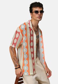 Koszula z krótkim rękawem, w odcieniach beżu, pomarańczy i niebieskiego, z siatkowym wzorem i geometrycznymi kształtami, noszona z jasnymi spodniami.