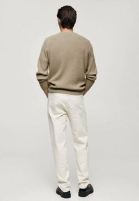 Bruine gebreide trui, witte broek en zwarte schoenen. De outfit heeft een gemêleerd patroon en een losse pasvorm, gezien vanuit de rug.
