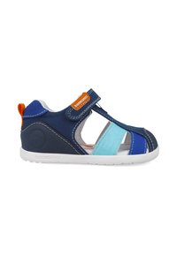Sandalia infantil en azul marino y azul con tiras elásticas y de Velcro, que presenta una etiqueta naranja. Parte superior de textil suave, suela blanca, diseño abierto para mayor transpirabilidad.