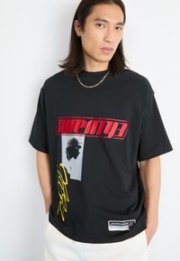 Black oversized t-shirt featuring red text, graphic image, and yellow accents. Includes a branded label at the bottom right. Cotton material.