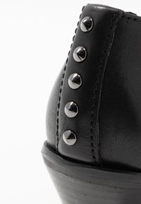 Bottine noire en cuir présentant un bout arrondi, une surface texturée et cinq clous métalliques sur le côté. Talon légèrement surélevé.